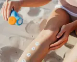 Main sur le sable, une main applique des points de crème solaire sur une jambe. Attention, cette crème solaire perimee pourrait ne plus protéger efficacement.