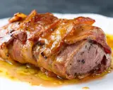 Saftiges Schweinefilet im Speckmantel, glasiert und mit Pfeffer bestreut, ruht in einer köstlichen Sauce auf einem weißen Teller.