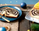 Tranches de roulé au pavot, garni de noix, sur une assiette bleue avec une fourchette dorée. Un autre roulé décoré d'orange est à droite.