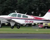 Eine weiße Beechcraft Bonanza mit roten und blauen Streifen hebt vom Gras ab.