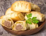 Stos pieczonego ciasta francuskiego z kiełbaską w środku, ozdobiony gałązką pietruszki.