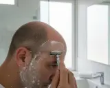 Mann rasiert sich die Glatze mit einem Rasierhobel und Rasierschaum im Badezimmer.