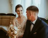 Uśmiechnięta panna młoda w sukni z koronkami i pan młody w granatowym garniturze. W tle kanapa i zasłony. Krótka ceremonia, bo ile trwa ślub cywilny?