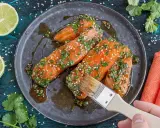Lachsfilets werden mit einer asiatischen Marinade aus Sojasauce, Ingwer, Knoblauch, Kräutern und Sesam bestrichen. Dazu Limettenspalten und Koriander.