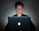 Młody mężczyzna pochylony nad laptopem, jego twarz oświetlona ekranem. Czy to początek uzależnienia od internetu?
