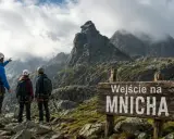 Trzech turystów w kaskach i z plecakami podziwia majestatyczne szczyty w Tatrach. Jeden z nich wskazuje drogę na Mnicha.