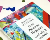 A book titled "How Painting Happens" by Martin Gayford, featuring a vibrant, abstract cover that evokes the spirit of figurative painting. Splatters of paint surround it.