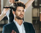Mężczyzna w eleganckim garniturze czeka na stylizację fryzury męskiej. Fryzjerka suszy jego włosy suszarką.