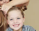 Ein Mädchen mit blauen Augen und Sommersprossen lächelt, während ihre Mutter ihr die Haare zu niedlichen Dutts frisiert. Tolle Ideen für kinder frisuren!