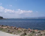 Słoneczny brzeg jak pić. Widok na morze i miasto w oddali, z kamienistym wybrzeżem na pierwszym planie.