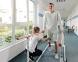 Młody mężczyzna ćwiczy chód przy poręczy, a fizjoterapeutka pomaga mu w rehabilitacji.