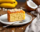 Tranche de banana bread moelleux, nappée de caramel et saupoudrée de noix de coco râpée. Des bananes et une noix de coco sont en arrière-plan.