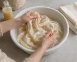 Manos lavando delicadamente una tela de seda color crema en un bol con agua jabonosa. Demostrando que la seda se puede lavar.