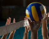 Piłka do siatkówki tuż nad siatką, gracze próbują ją odbić. Widać ich dłonie i fragmenty siatki, która wyznacza wysokość siatki w siatkówce.