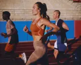 Biegacze na bieżni. Kobieta w pomarańczowym topie i beżowych legginsach, mężczyzna w koszulce i szortach. Buty sportowe z niskim dropem zapewniają komfort.