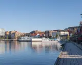 En vacker dag att besöka Luleå. En träspång leder längs vattnet förbi röda byggnader och en vit båt.