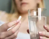 Dłoń trzymająca białą kapsułkę i szklankę wody.