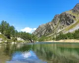 Il **lago d'Arpy** riflette il cielo azzurro e le montagne rocciose. Sulle rive, pini e persone godono del sole estivo.
