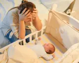 Madre agotada, con las manos en la cabeza, mira a su bebe que se desvela por la noche en su cuna.