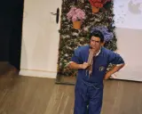 Actor con mono azul y pañuelo a cuadros, actuando en un teatro para niños. Fondo con flores y enredaderas.