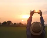 Osoba w kapeluszu fotografuje zachód słońca telefonem z aparatem szerokokątnym.