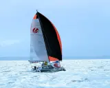 Voilier Heol 7.4 fendant les vagues, ses voiles bicolores gonflées par le vent. Deux marins à bord.
