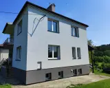 Jasny dom typu kostka z szarym cokołem i balkonem, otoczony zielenią.