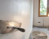 Narzędzie do wygładzania ścian, pokazujące jak położyć gładź. W tle wiadro z masą i okno.