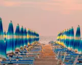 Filetto di ombrelloni a righe blu e bianche sulla spiaggia, con il mare e il cielo rosa all'orizzonte. Un'immagine iconica della riviera adriatica.