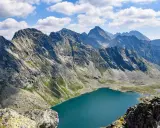 Widok na turkusowe jezioro u podnóża skalistych gór. W oddali widać kolejne pasma górskie, a na niebie chmury. To właśnie Koprowy Wierch.