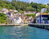 Bunte Häuser schmiegen sich an einen grünen Hang in Cudillero, Spanien. Das türkisfarbene Wasser des Hafens spiegelt die malerische Kulisse wider.