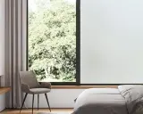 Habitación con cama, sillón y ventana con vidrio mate que filtra la luz del sol y la vegetación exterior.