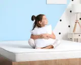 Niña sonriente abraza almohada en un colchón, pensando qué colchon es mejor para niños.