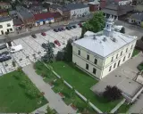 Sławków rynek z lotu ptaka: żółty budynek ratusza, brukowany plac z samochodami i zielony park z ławkami.