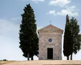 Piccola chiesa in pietra con cipressi, icona del Val d'Orcia, sotto un cielo azzurro con nuvole bianche.