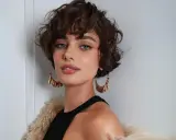 Mujer con un moderno corte shaggy rizado, ojos azules penetrantes y grandes pendientes dorados.
