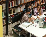 Trzy studentki pracują przy stole w bibliotece wyższej szkoły technicznej, otoczone książkami i laptopem.
