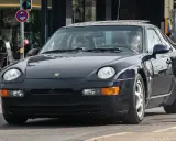 Dunkelblauer 968 Porsche fährt auf der Straße. Ein Mann sitzt am Steuer. Verkehrsschilder sind im Hintergrund zu sehen.