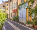 Bunte Häuser und Pflanzen säumen eine Gasse in der Marseille Altstadt. Ein "Bazar du Panier" Schild und blaue Holztüren fallen auf.