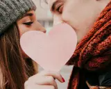 Para zakochanych, ukrywająca się za różową kartą w kształcie serca. Ich twarze są blisko siebie, tworząc intymną scenę.