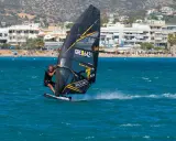 Windsurfer mit schwarzem Segel und gelben Akzenten auf türkisfarbenem Wasser vor einer Küste mit Gebäuden und Bergen. Surfen in Griechenland.