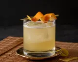 Ein erfrischender Rum Sour mit Eis und Orangenzesten garniert, serviert auf einem Bambusuntersetzer.