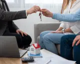 Agente inmobiliario entrega llaves a pareja. Un modelo de casa y calculadora sugieren una transacción, no una notificación de lanzamiento de desahucio.