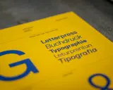 Żółta kartka z niebieskimi napisami: Letterpress, Buchdruck, Typographie, Leturpretun, Tipografia. Duża litera "G" po lewej. To praca typografa.