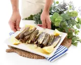 Manos sirviendo un plato de navajas a la plancha, decorado con gajos de limón y perejil picado.