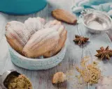 Un bol de biscuits madeleine noisette saupoudrés de sucre glace, avec des étoiles de badiane et une cuillère de poudre sur une table en bois.