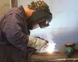 Soldador trabajando con un electrodo, creando chispas y humo. Se aprecian partes del electrodo en acción.