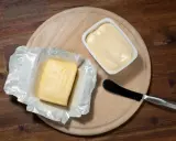 Zwei Butterpackungen, eine in Folie, die andere in einer weißen Schale, liegen auf einem Holzbrett. Eine leichte Butter weniger Fett für den gesunden Genuss.