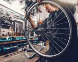 Mechanik sprawdza układy w rowerze, trzymając koło. W tle warsztat z narzędziami.