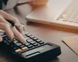 Osoba liczy na kalkulatorze, zastanawiając się, czy studia stacjonarne są płatne. Obok leży laptop i koperta.